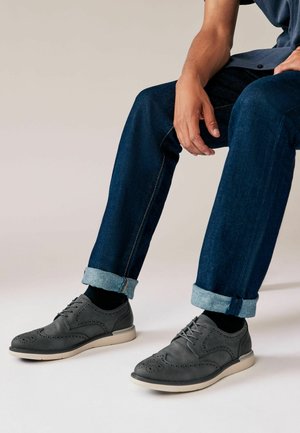 Chaussures de ville en cuir gris avec des accents perforés, associées à un jean en denim bleu retroussé. Les chaussures présentent une semelle de couleur claire et des lacets.
