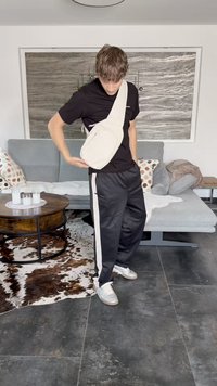 Beige crossbody bag made of soft fabric, rounded shape, zipper closure, worn on the shoulder. Black t-shirt and track pants with white stripes.