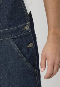 Salopette en denim bleu foncé avec une poche latérale, des accents de boutons métalliques et des coutures jaunes pour le contraste. Texture lisse, design fuselé.