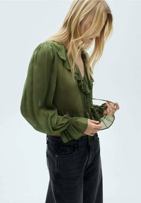 Blonde Frau, die die Bänder einer durchsichtigen olivgrünen Bluse mit rüschigem Kragen und Ballonärmeln anpasst, in schwarzen hoch taillierten Jeans.