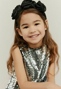 Lesklé strieborné šaty so flitrami bez rukávov, doplnené čiernym kvetinovým čelenkom z látky. Hladké vlasy a svetlý tón pleti.