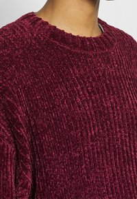 Bordový sametový svetr s žebrovanou strukturou, kulatým výstřihem a jemným leskem. Detailní záběr na látku a šití.