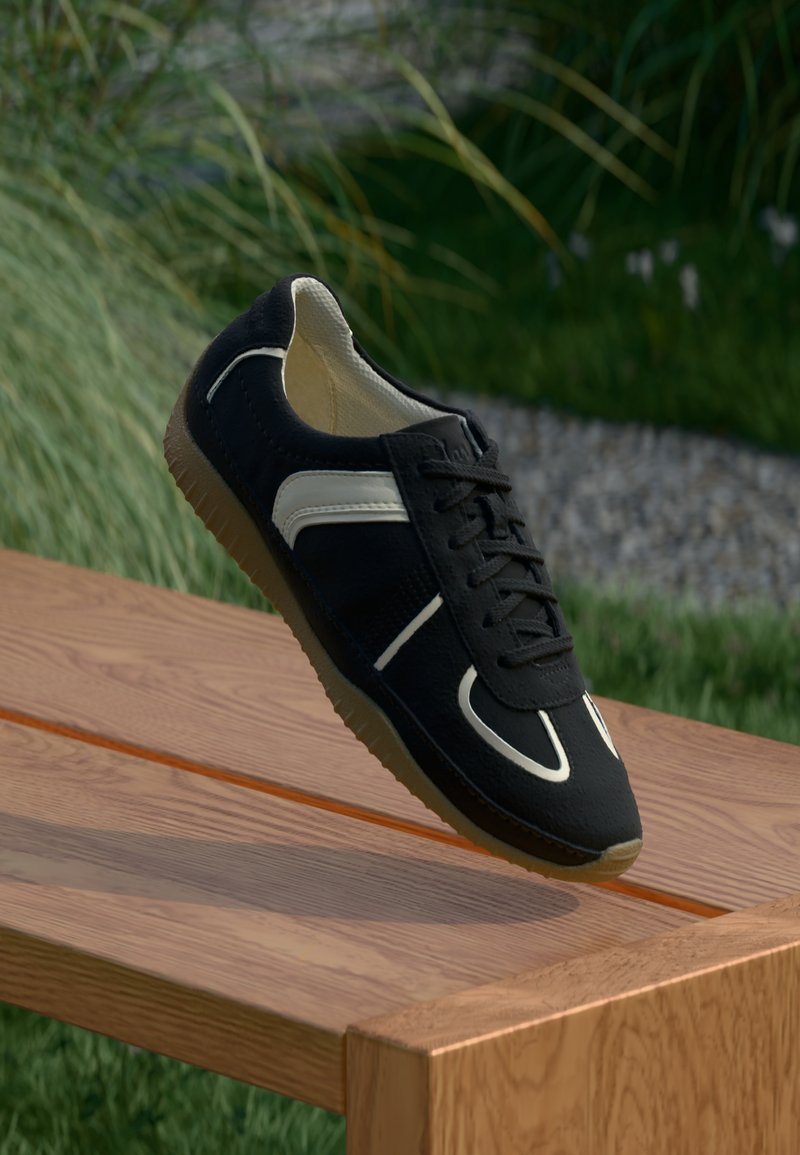 Svart-hvite sneakers med gummisåle som hviler på kanten av en trebenk ute, med gress og planter i bakgrunnen.