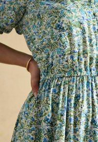 Krupni plan osobe u plavoj, zelenoj i bijeloj cvjetnoj haljini s povećanim strukom, ruka joj je na kuku, a na zapešću nosi zlatnu narukvicu.