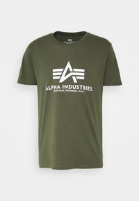 Olivově zelené bavlněné tričko s centrálním bílým grafickým logem s nápisem "ALPHA INDUSTRIES" a třemi horizontálními pruhy pod ním.