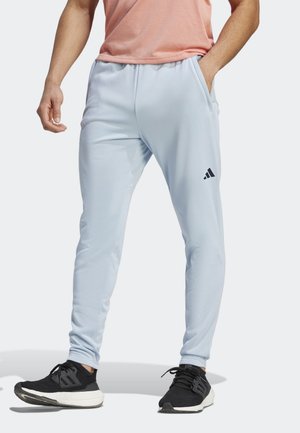 Man die lichtblauwe Adidas joggingbroek, zwarte sneakers en een perzikkleurig T-shirt draagt, staand met één hand in de zak tegen een witte achtergrond.