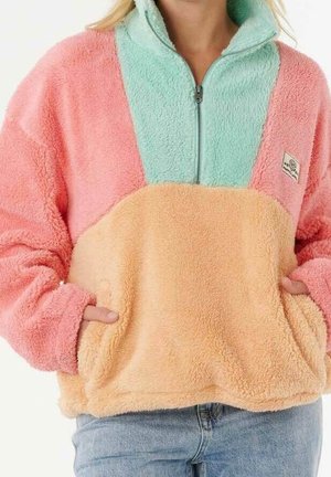 Woman wearing a pastel fleece pullover with pink sleeves, mint green collar, peach body, front zipper, and hands in front pockets.