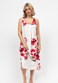 Donna con capelli biondi alla spalla che indossa un vestito bianco senza maniche con grandi stampe floreali rosse e rosa, in piedi a piedi nudi.