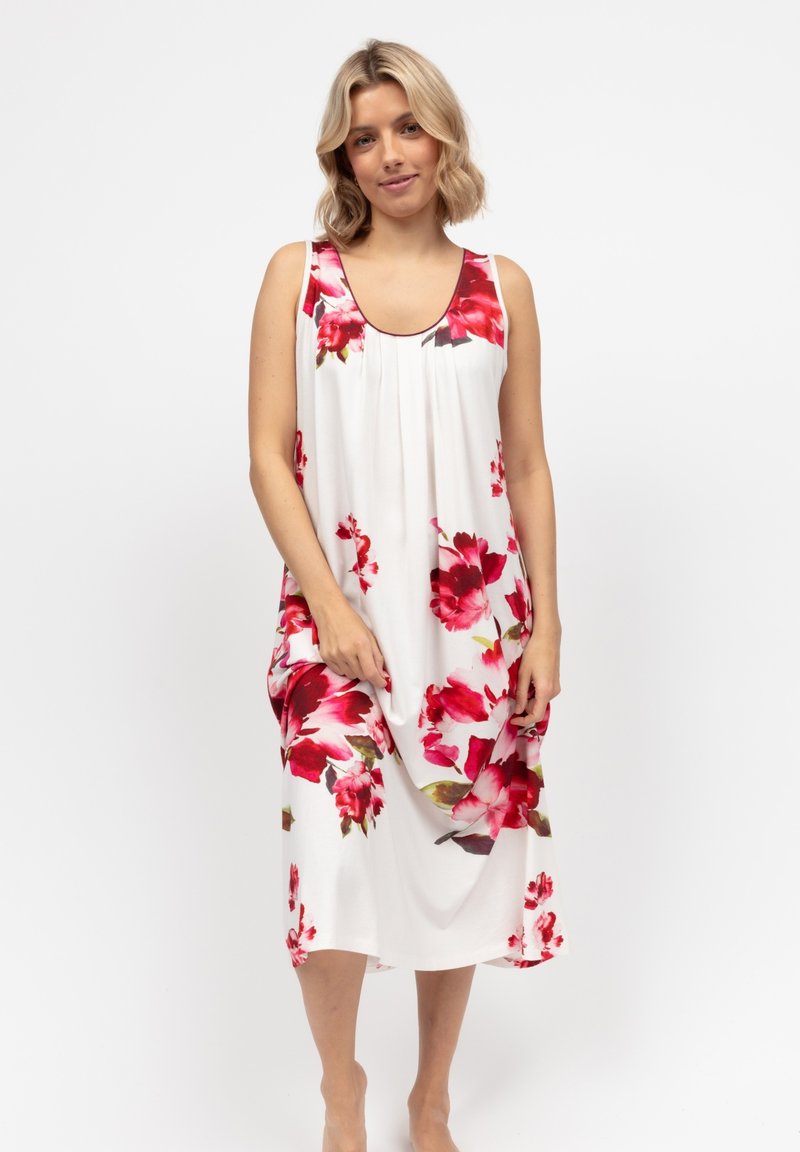 Donna con capelli biondi alla spalla che indossa un vestito bianco senza maniche con grandi stampe floreali rosse e rosa, in piedi a piedi nudi.