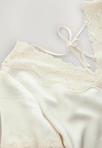 Caraco en soie crème avec bordure en dentelle et fines bretelles réglables nouées en nœud sur un fond clair.