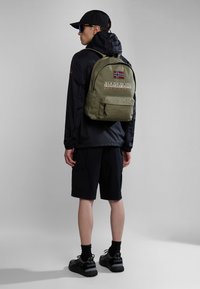 Olivově zelený batoh s přední kapsou, logo "NAPAPIJRI" a detail norské vlajky. Model má na sobě černou bundu, šortky a boty.