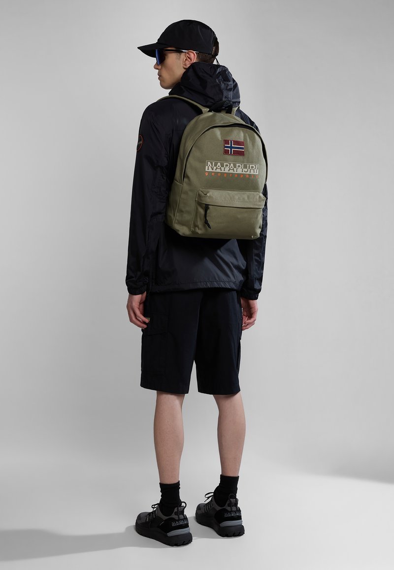 Olivově zelený batoh s přední kapsou, logo "NAPAPIJRI" a detail norské vlajky. Model má na sobě černou bundu, šortky a boty.