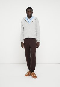 Grauer Zip-Hoodie mit einer Fronttasche, über einem blau-weiß gestreiften Shirt getragen. Braune Kordelhosen und sandfarbene Sneaker vervollständigen den Look.