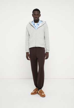 Grijze zip-up hoodie met een voorzak, gelaagd over een blauw-witte gestreepte shirt. Bruine broek met koord en tan sneakers maken de outfit compleet.