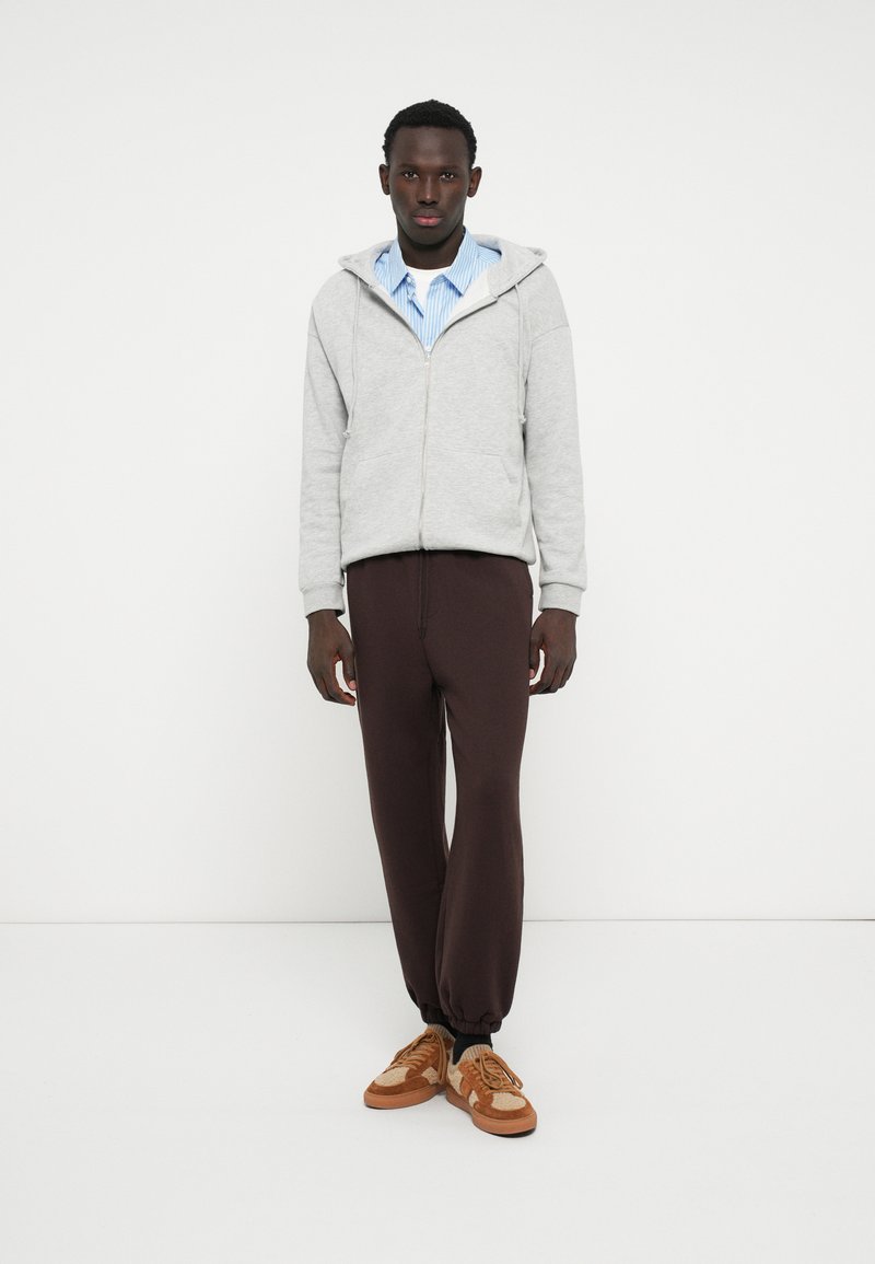 Grauer Zip-Hoodie mit einer Fronttasche, über einem blau-weiß gestreiften Shirt getragen. Braune Kordelhosen und sandfarbene Sneaker vervollständigen den Look.