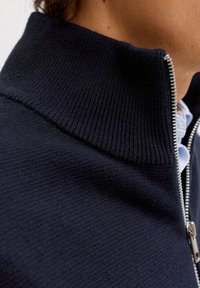 Μπλε ζακέτα με φερμουάρ, που διαθέτει ριμπ κολάρο και πλεκτή υφή. Η ασημένια λεπτομέρεια του φερμουάρ προσθέτει αντίθεση. Η κοντινή λήψη τονίζει τη δομή του υφάσματος.