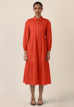 Frau steht und trägt ein langärmliges, geknöpftes, mit Kragen versehenes, gestuftes Midikleid in leuchtendem Orange mit schwarzen offenen Sandalen vor einem schlichten Hintergrund.
