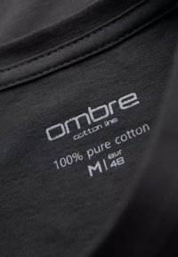 Sort bomulds T-shirt-mærkat med påskriften "ombre cotton line," "100% ren bomuld," og størrelsesoplysninger (M, EUR 48) i hvid tekst.