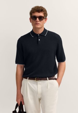Mann trägt Schildpatt-Sonnenbrille, schwarzen, kurzärmeligen, gerippten Polo, weiße Hose, braunen geflochtenen Gürtel, hält eine schwarze Tasche in der Hand.