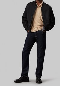 Veste bomber noire, polo beige, jeans bleu marine et chaussures noires. La veste dispose d'une fermeture éclair et de poches latérales. Textures polies et lisses.