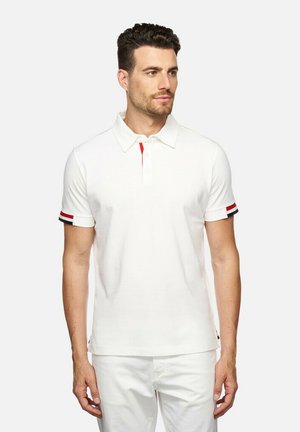 Camisa polo blanca con una tapeta de tres botones, acento rojo debajo del cuello y puños a rayas en rojo, blanco y azul. Mangas cortas.