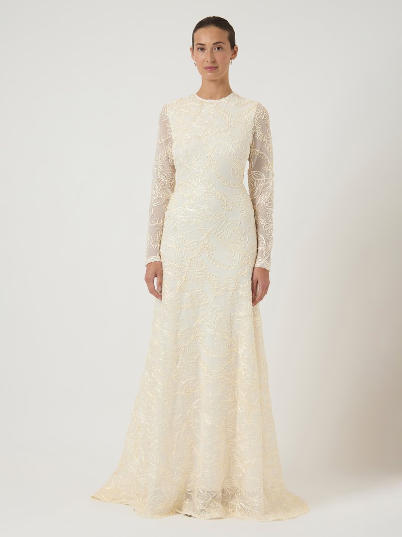 Γυναίκα με μακριά μανίκια και μακρύ λευκό δαντελένιο φόρεμα με λουλουδάτη κέντημα, που στέκεται σε απλό λευκό φόντο.