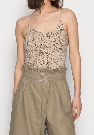 Femme portant un top beige à fines bretelles avec des pois noirs et un pantalon ample kaki taille haute avec ceinture et taille froncée.