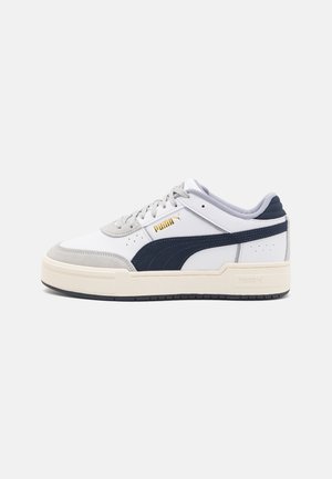 Witte lage sneaker met donkerblauwe Puma-streep, beige zool, grijze suède neuskap en gouden Puma-logo aan de zijkant.