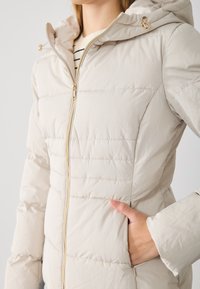 Doudoune beige avec fermeture éclair devant, capuche et poches latérales. Présente une texture matelassée horizontale et des accents en métal doré.