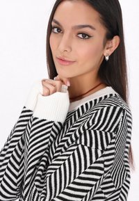 Pulover cu dungi alb-negru, cu un model în zig-zag, manșete cu striații și o textură moale. Modelul are părul lung și brunet și poartă cercei din argint.