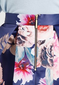 Blommönstrad tyg med stora rosa, lila och beige blommor och blad, med en synlig guldfärgad dragkedja i mitten.