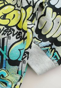 Bluza z szarym i czarnym wzorem graficznym z żółtymi i niebieskimi akcentami. Teksturowana tkanina z ściągaczami przy mankietach i szwach.