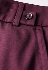 Tessuto bordeaux con una superficie liscia, dotato di una chiusura con un solo bottone e passante per cintura. La cucitura lungo le cuciture è visibile.