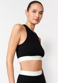 Czarny sportowy crop top z okrągłym dekoltem i białą elastyczną taśmą z logo na dole. Gładka faktura, dopasowany krój.