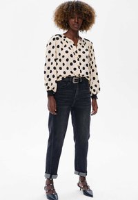 Kobieta z kręconymi włosami, ubrana w beżową bluzkę z dużymi czarnymi kropkami, czarne dżinsy z wysokim stanem oraz czarne sandały na obcasach z perłami.