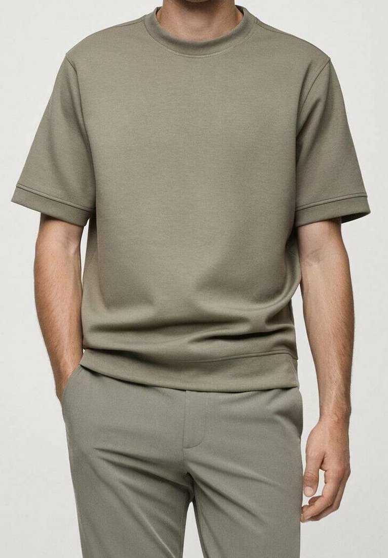 Homme portant un t-shirt vert olive clair à manches courtes et un pantalon assorti, une main dans la poche, sur un fond uni.