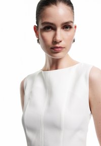 Vestido blanco sin mangas con un diseño estructurado, que presenta detalles de costura sutiles y una textura suave. Modelo que lleva pendientes de aro de plata.