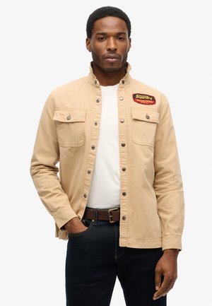 Beżowa bawełniana kurtka z zatrzaskami, dwoma kieszeniami na piersi i czerwoną naszywką "Superdry Industries". Nosi ją na białej koszuli z ciemnoniebieskimi dżinsami.