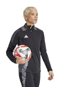 Veste de sport noire avec accents gris, fermeture éclair complète et design ajusté. Une personne tient un ballon de soccer à motifs rouge et blanc.