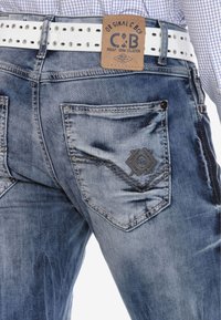 Vervaagde blauwe spijkerbroeken met gestikt patroon op de zakken, een bruine leren patch en een witte riem met metalen oogjes.