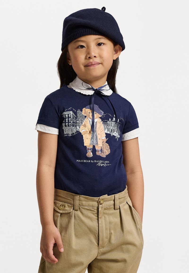 Ναυτικό βαμβακερό T-shirt με λευκό γιακά, που διαθέτει γραφικό με αρκούδα σε μπεζ παλτό. Ταμπά, πλυμένη παντελόνα με λεπτομέρειες από κουμπιά.