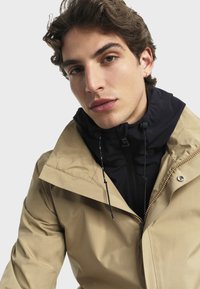 Beige Außensakko mit einer schwarzen Innenschicht, Reißverschluss und Kordelzugkragen. Glatte Textur und minimalistisches Design mit sichtbaren Beschlägen.