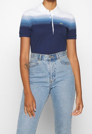 Vrouw draagt een kortärmelig polo-shirt met blauw-witte strepen en een lichte, hooggetailleerde jeans, met een tatoeage op haar linker onderarm.