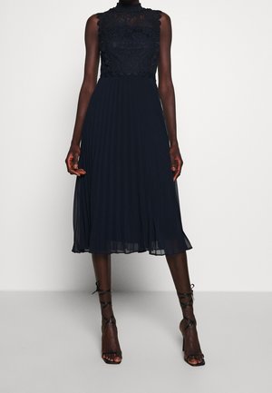 Tummansininen laskostettu mekko pitsipuvun kanssa ja korkea pääntie. Ominaisuuksiin kuuluu monimutkainen pitsikoristelu ja levenevä hame, joka ulottuu polven alapuolelle.