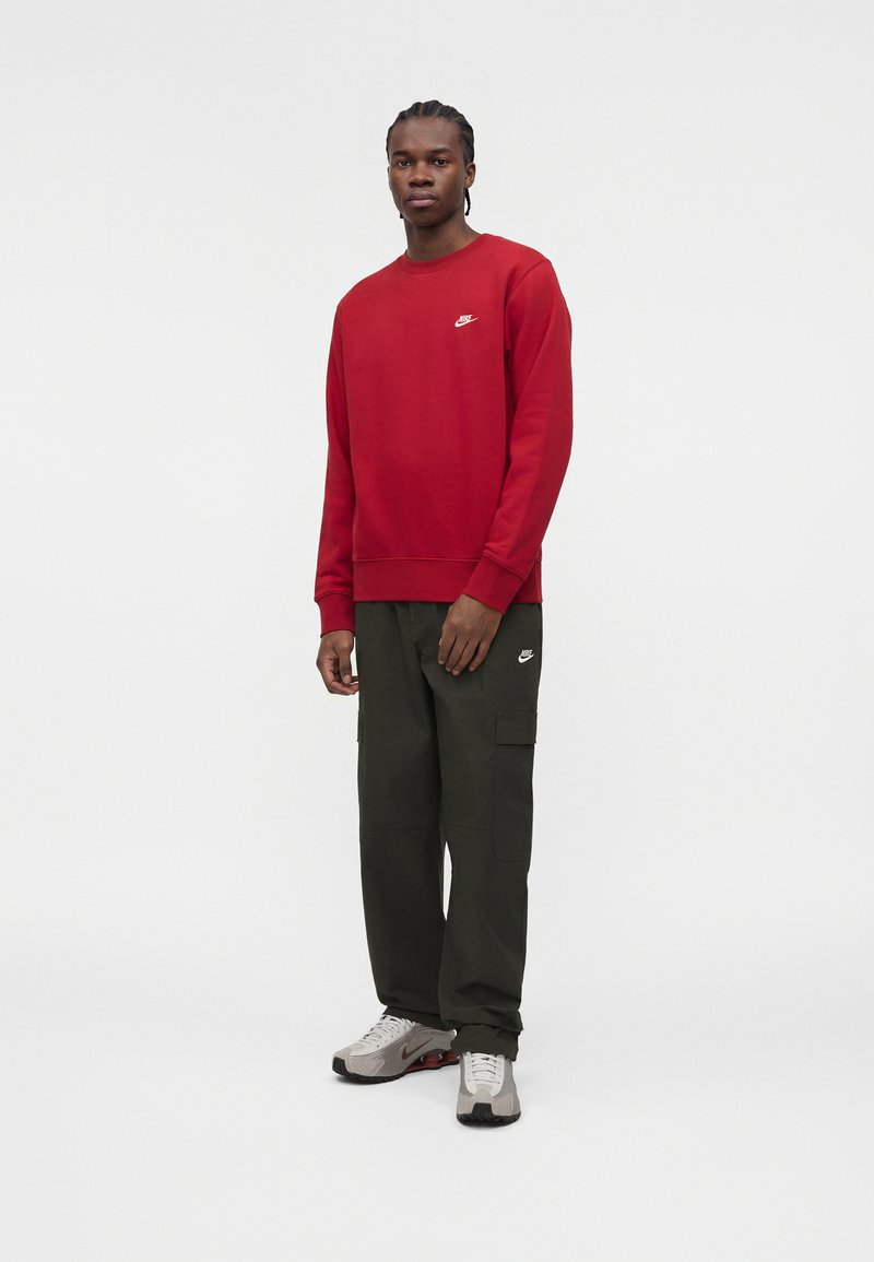 Hombre de pie con sudadera roja de Nike, pantalones cargo oscuros y zapatillas Nike grises sobre fondo blanco liso.