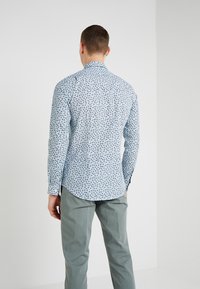 Camicia a maniche lunghe da uomo in tessuto azzurro chiaro con un motivo floreale. Presenta un orlo curvo e dettagli con polsini abbottonati. Abbinata a pantaloni verde chiaro.