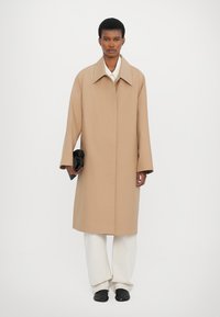 Beige long coat with a wide collar and straight silhouette. Model holds a black clutch. Worn over white pants and black shoes.