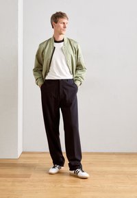 Un homme se tient avec les mains dans les poches, portant une veste bomber vert clair, un t-shirt à côtes blanc, un pantalon noir et des baskets blanches avec des rayures noires.