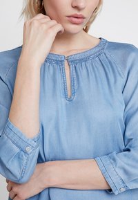 Ljusblå chambrayskjorta med nyckelhålsknäppning, inrullat detalj och 3/4 ärmar med knäpplångd. Slät, något strukturerad tyg.