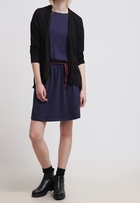 Czarny kardigan na granatowej sukience z bordowym paskiem. Modelka ma na sobie czarne botki. Gładki materiał; prosty, casualowy design.